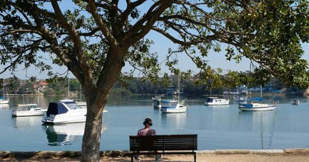 A persons sitting on a bench under a tree looking over the water view at Kind George Park.